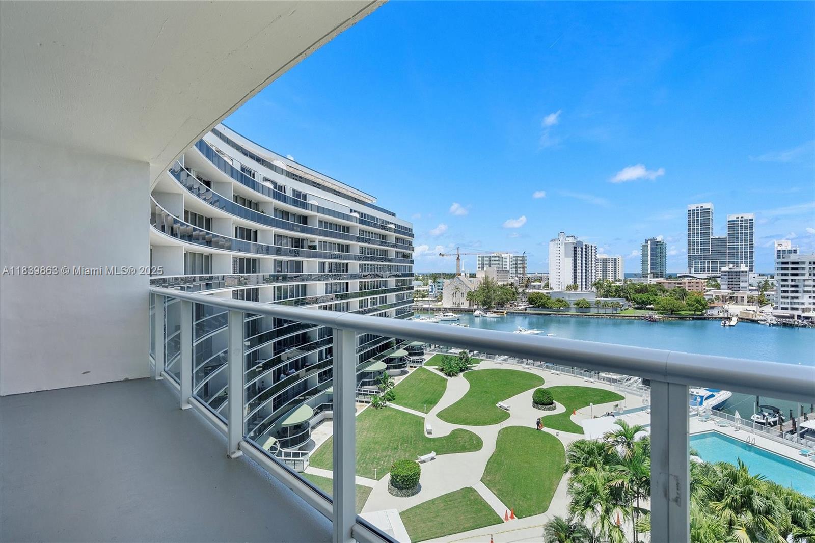 A balcony overlooks a curved apartment building, a landscaped park, a waterway, and city skyline — 900 Bay Dr, Miami Beach FL 33141, MIAMI BEACH, FL