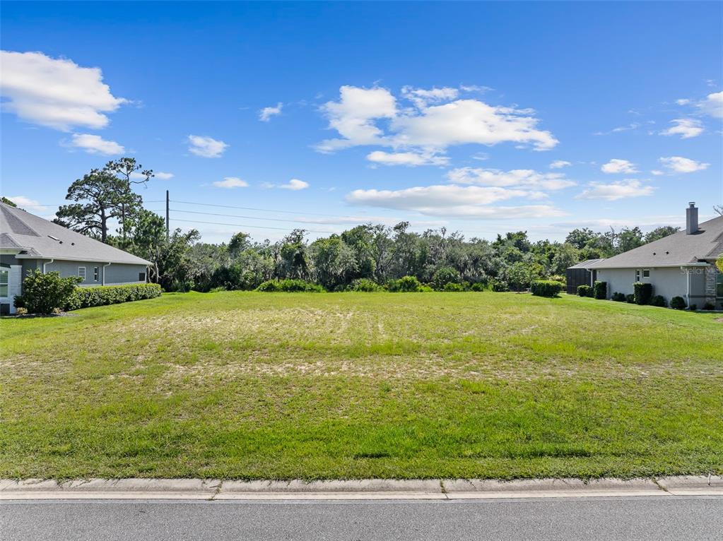 A vacant, grassy residential lot sits between two houses under a blue sky — 3040 ACOMA DRIVE, ORMOND BEACH, FL