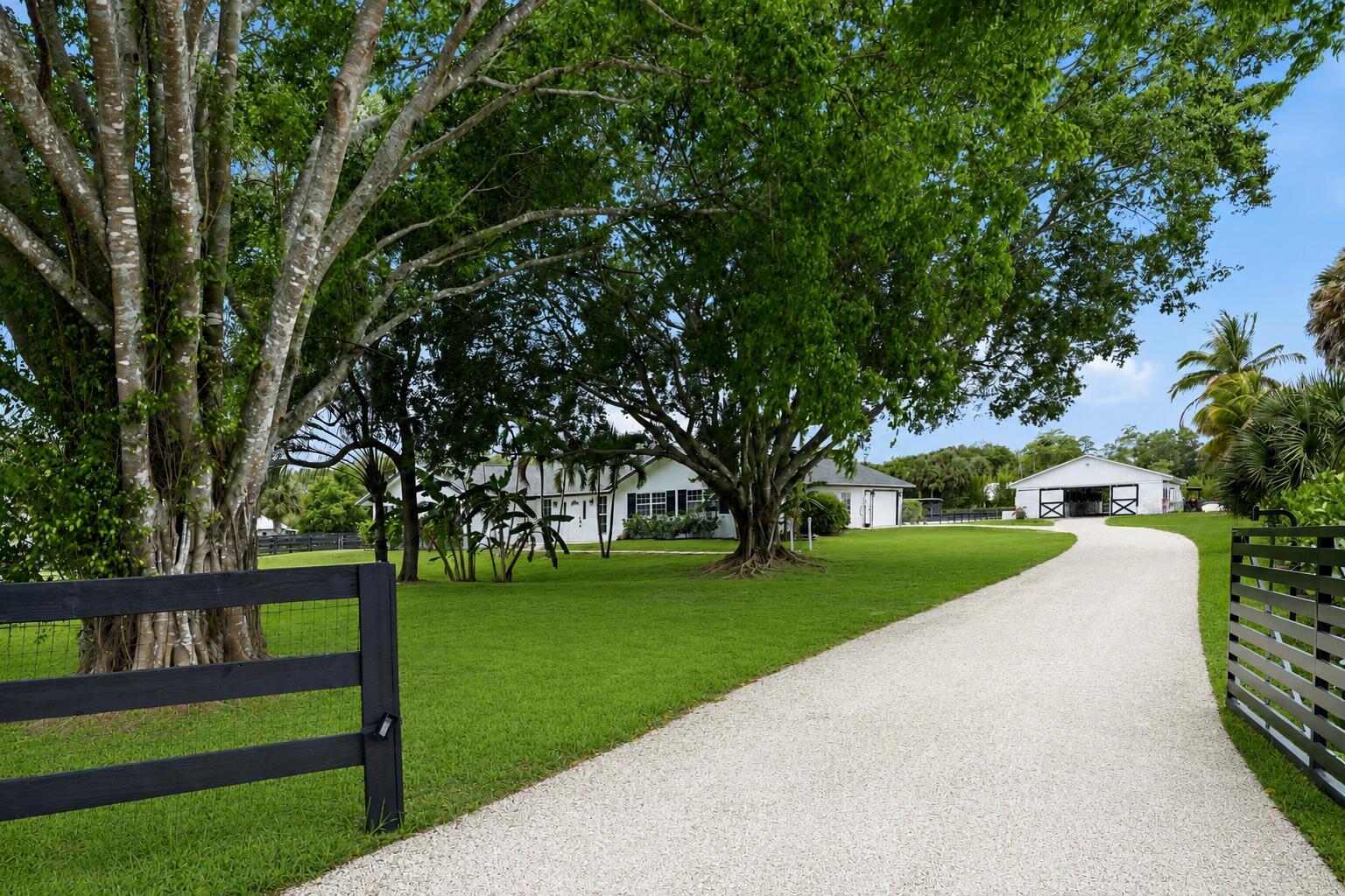 A gravel driveway leads to a white house and barn surrounded by large green trees — 785 Cindy Drive, Wellington FL 33414, WELLINGTON, FL