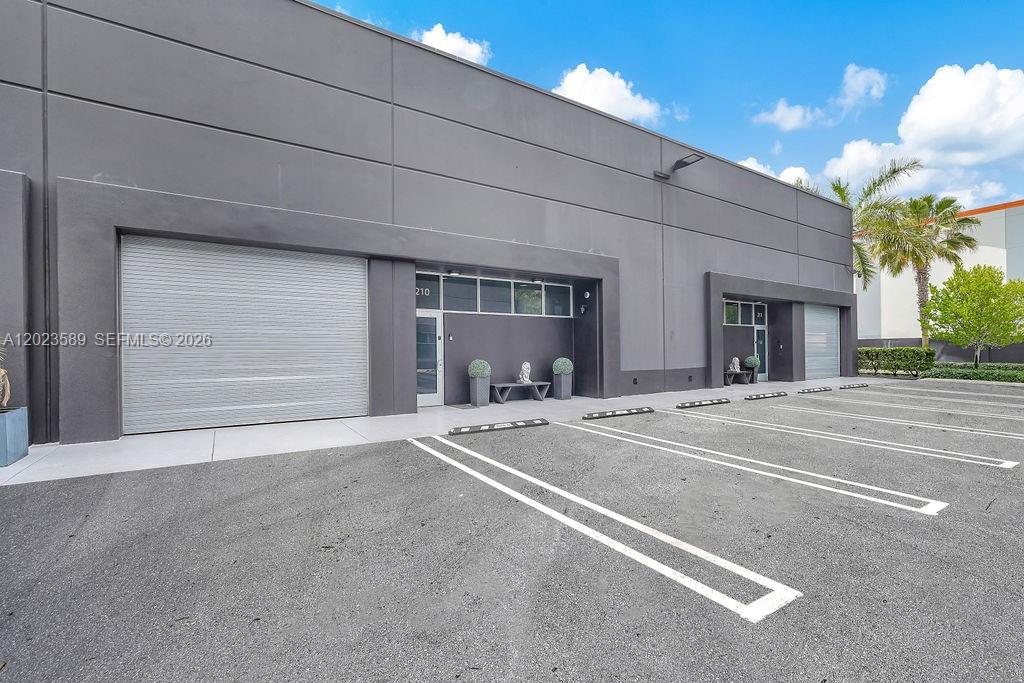 The exterior of a modern commercial building features gray walls, roll-up doors, and parking spaces — 6030 NW 102nd Ave # 210, Doral FL 33178, DORAL, FL