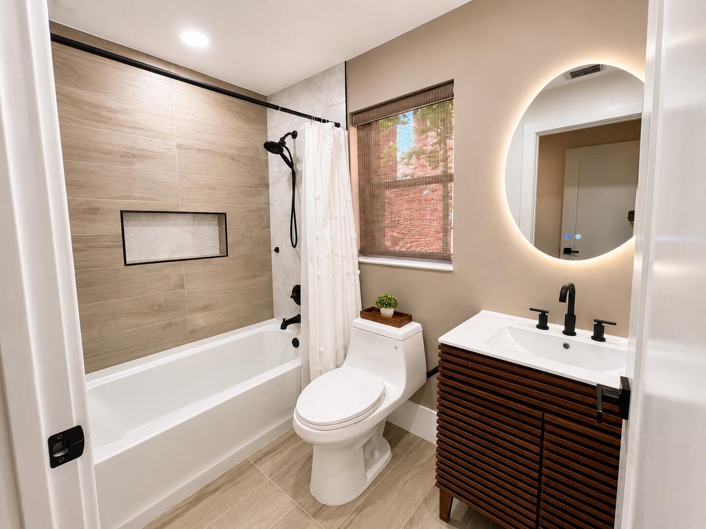 A modern bathroom features a wood-look tile tub surround, a slatted vanity, and a round mirror — 1073 Aspri Way, Riviera Beach FL 33418, RIVIERA BEACH, FL