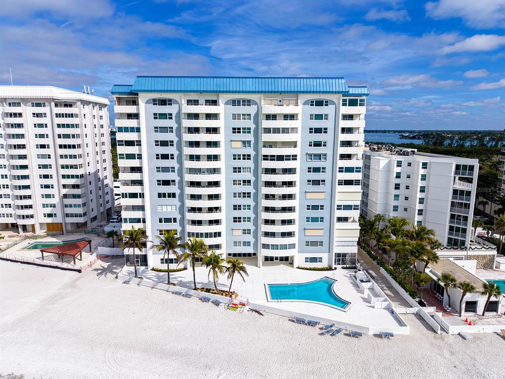 An aerial view shows a multi-story beachfront condominium building with a pool and palm trees — 1750 BENJAMIN FRANKLIN DRIVE UNIT 9F, SARASOTA, FL