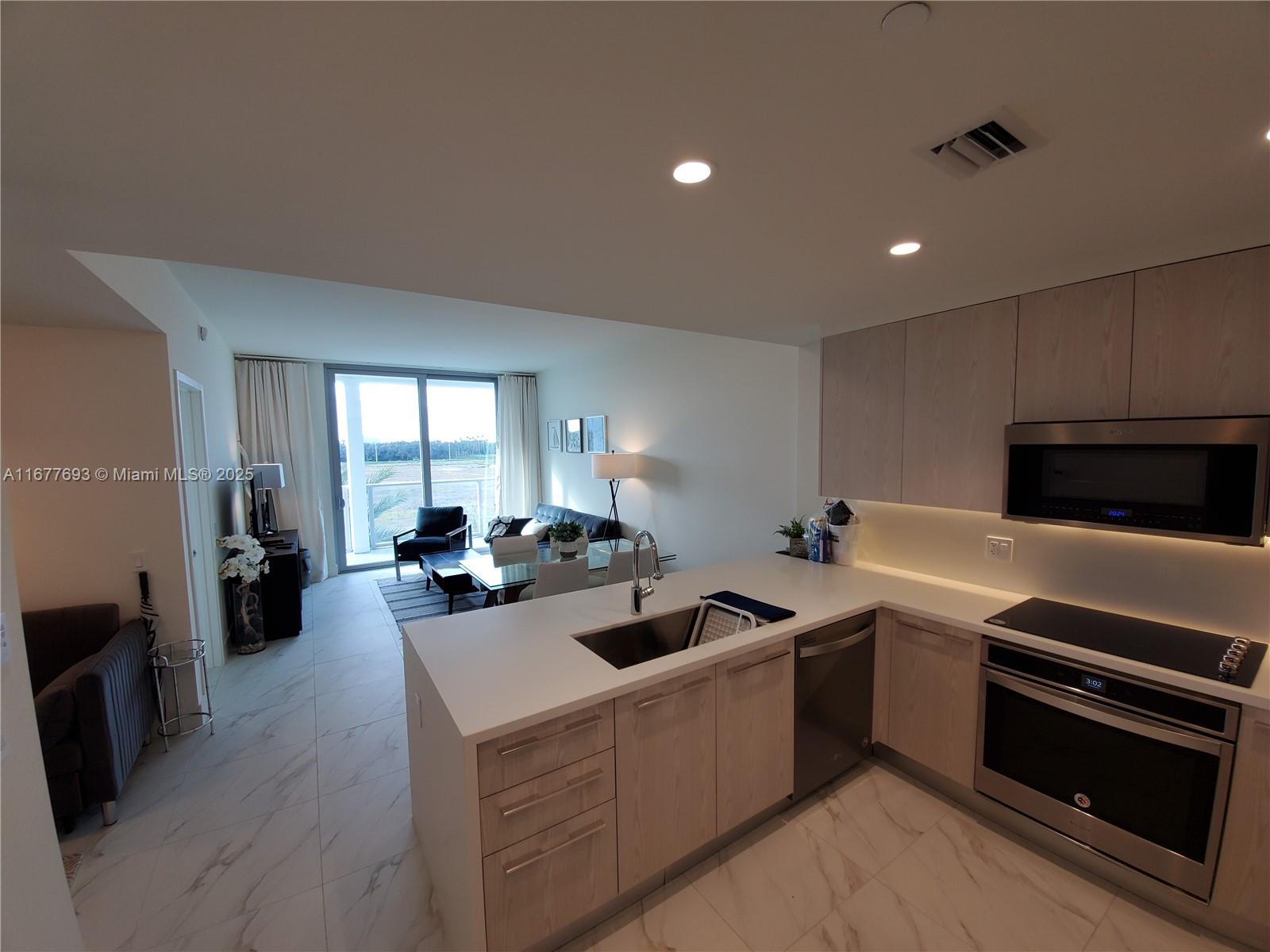 A modern kitchen with light wood cabinets and white countertops overlooks an open-concept living area — 2000 Metropica Way # 305, Sunrise FL 33323, SUNRISE, FL
