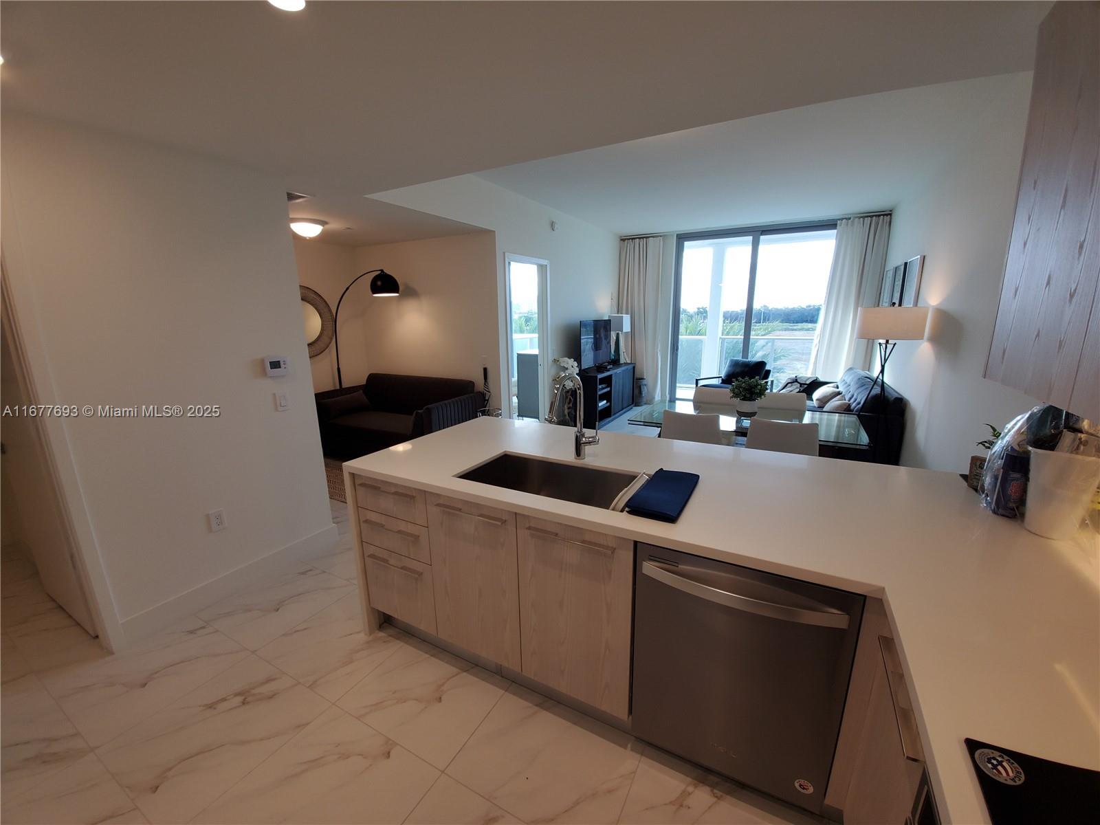 A modern kitchen with light wood cabinets and white countertops overlooks an open living area — 2000 Metropica Way # 305, Sunrise FL 33323, SUNRISE, FL