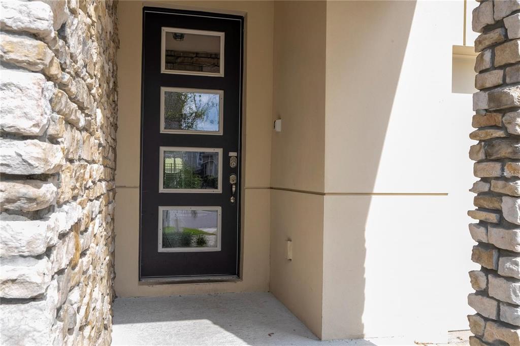 A modern dark-framed front door with four glass panels sits between two stone-clad walls — 9043 SUNSHINE RIDGE LOOP, KISSIMMEE, FL