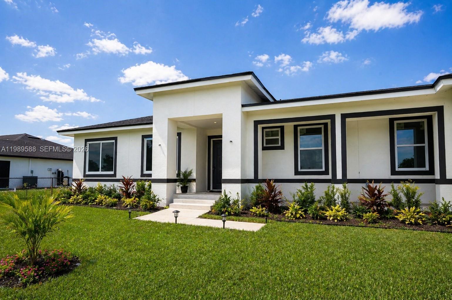 A single-story white house with dark trim features a central entryway and landscaped front yard — 1715 McArthur Ave, Lehigh Acres FL 33972, LEHIGH ACRES, FL