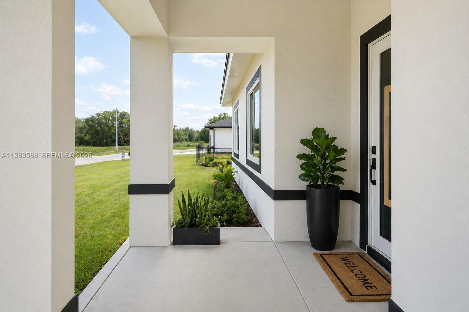A covered concrete porch features a potted plant, a welcome mat, and white walls — 1715 McArthur Ave, Lehigh Acres FL 33972, LEHIGH ACRES, FL