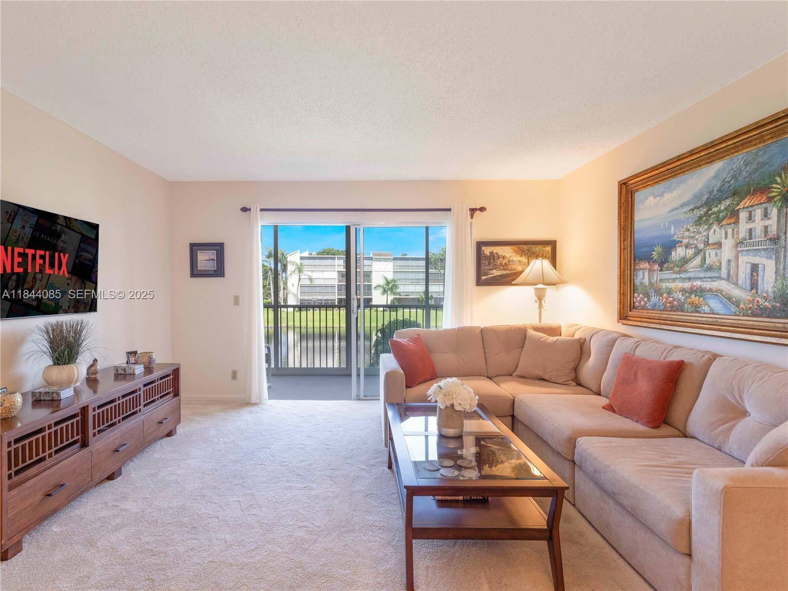 A living room features a beige sectional sofa, a wooden media console, and a balcony view — 14360 Strathmore Lane # 203, Delray Beach FL 33446, DELRAY BEACH, FL