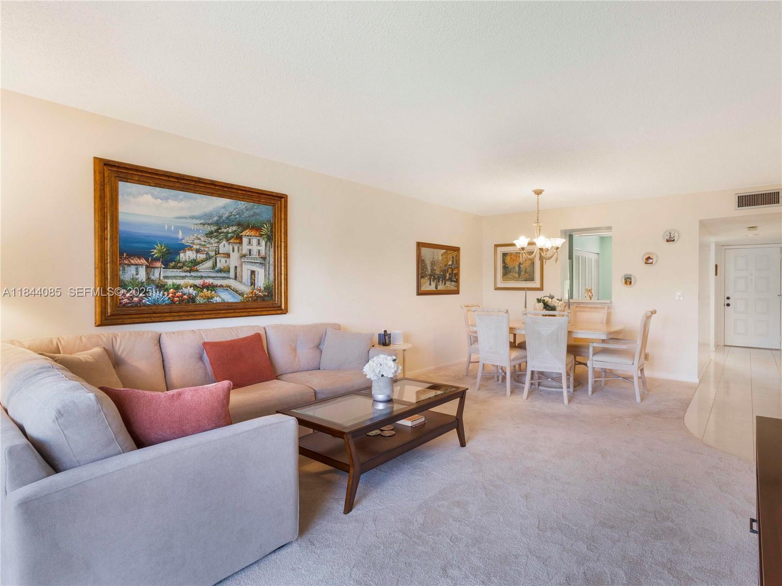 A living room and dining area feature beige carpeting, a sectional sofa, and wall art — 14360 Strathmore Lane # 203, Delray Beach FL 33446, DELRAY BEACH, FL