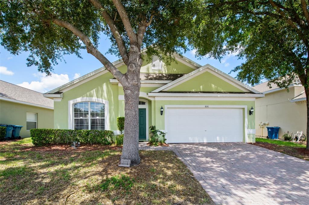 A light green single-story house with a two-car garage and a large tree in front — 826 ANDALUSIA LOOP, DAVENPORT, FL