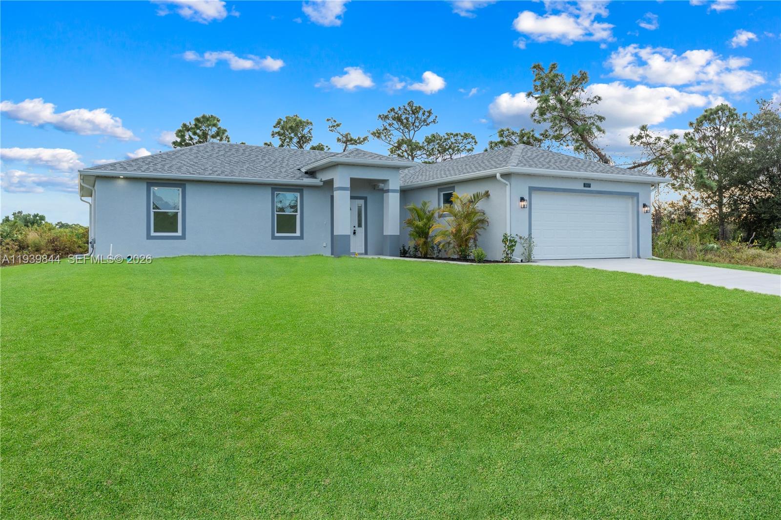 A single-story gray house with a two-car garage sits behind a large green lawn — 3117 W 64th W, Lehigh Acres FL 33971, LEHIGH ACRES, FL