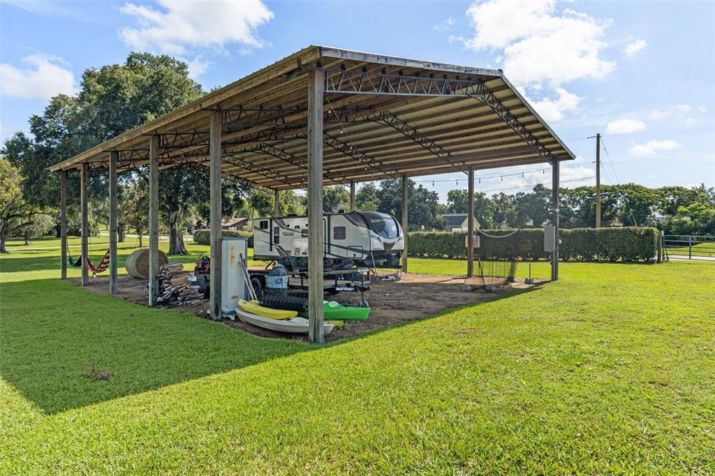 A large metal-roofed carport shelters a travel trailer, kayaks, and firewood on a grassy lot — 37714 FAIRFIELD LANE, DADE CITY, FL