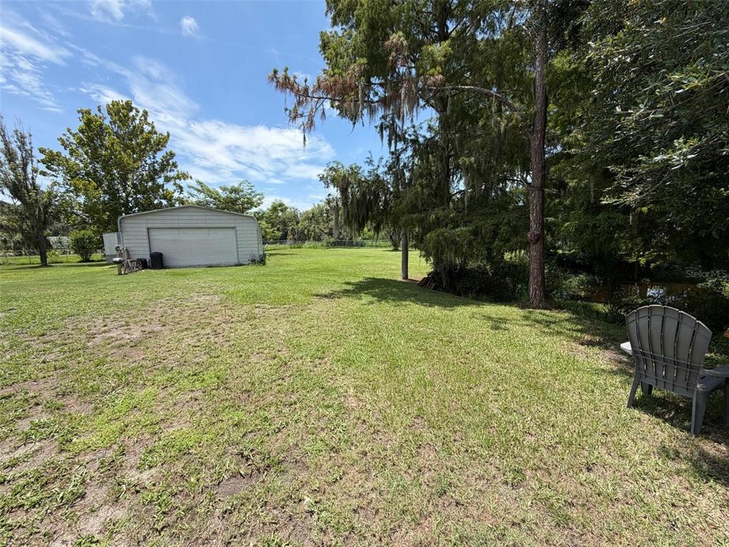 A grassy backyard features a detached metal garage, mature trees, and a plastic chair — 3707 36TH STREET E, BRADENTON, FL