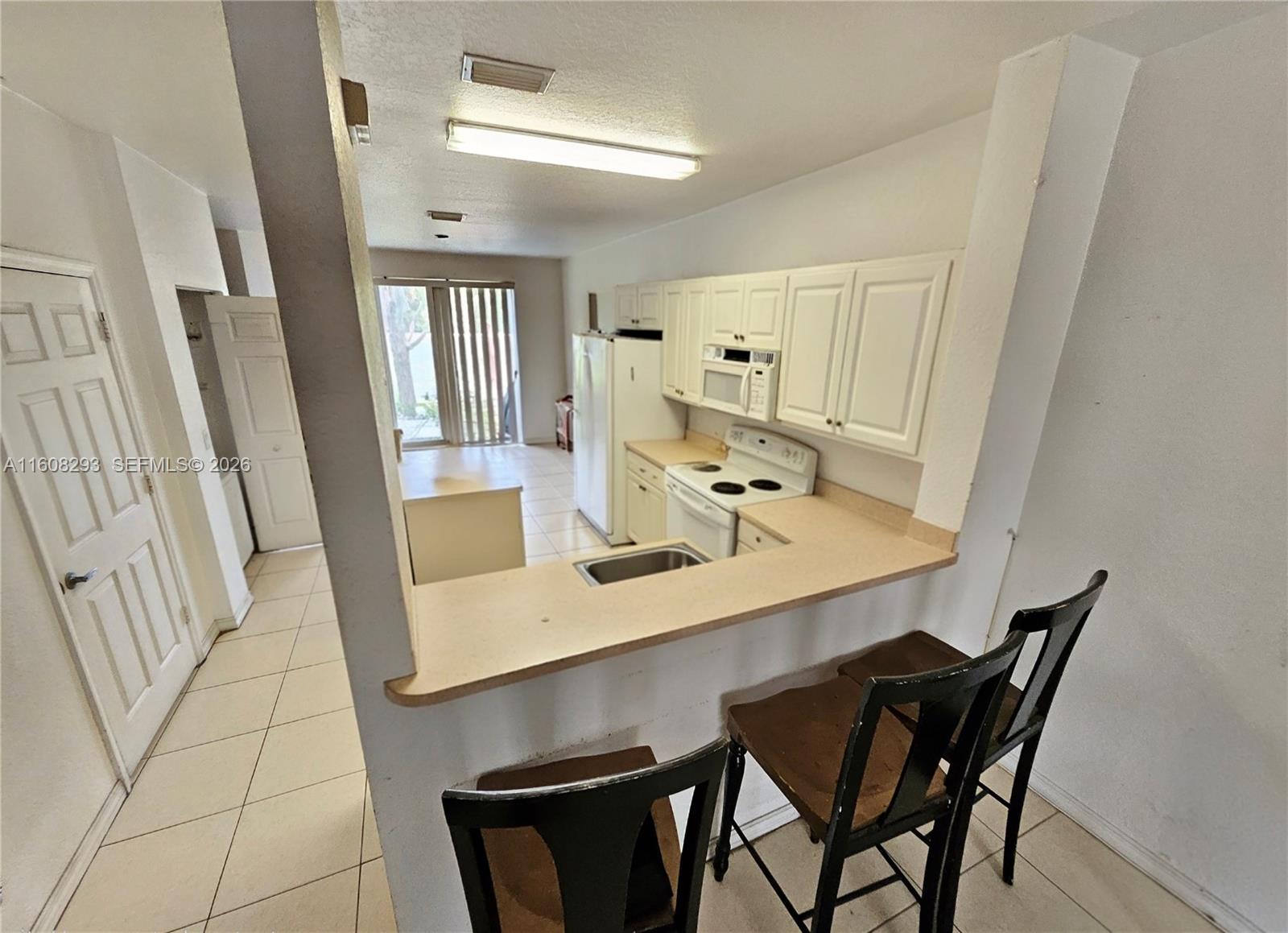 A kitchen with white cabinets, a breakfast bar, and tile flooring overlooks a living area — 3556 Oleander Ter # 3556, Riviera Beach FL 33404, RIVIERA BEACH, FL