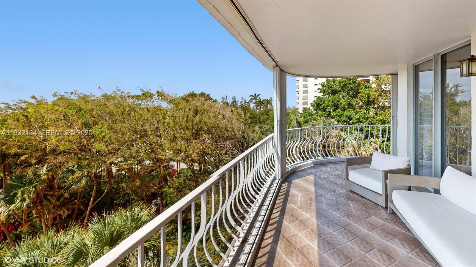 A curved balcony features tiled flooring, white outdoor furniture, and a metal railing overlooking trees — 10155 Collins Ave # 410, Bal Harbour FL 33154, BAL HARBOUR, FL