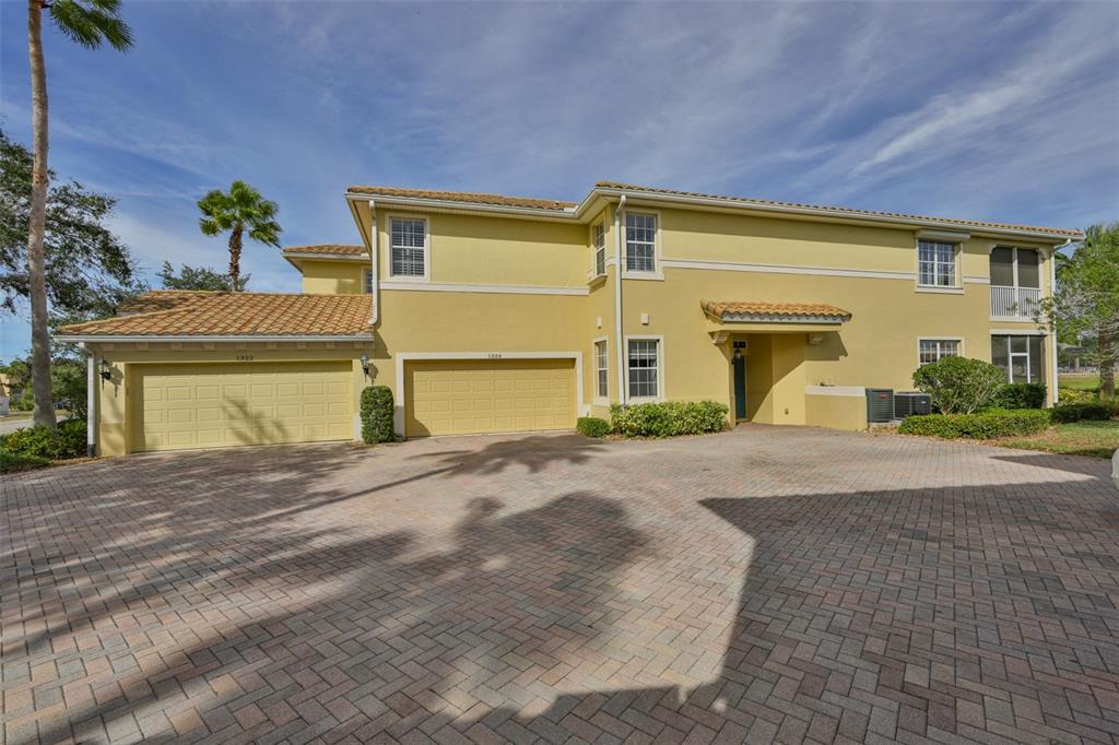 A two-story yellow stucco house features a paver driveway and two attached garage doors — 1324 EMERALD DUNES DRIVE, SUN CITY CENTER, FL