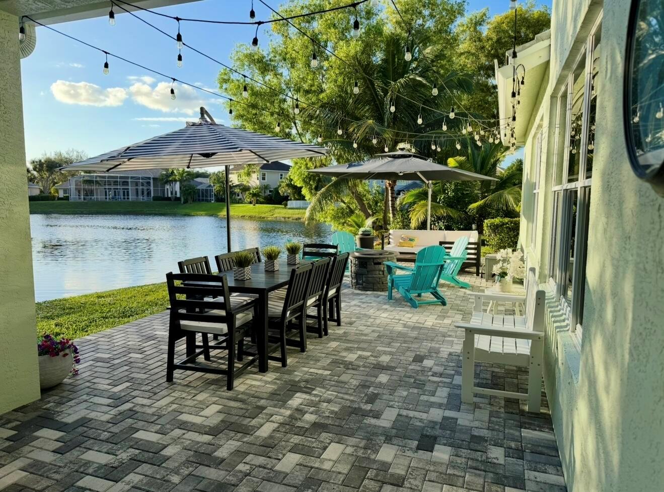 A paved patio overlooks a lake with outdoor dining, seating, umbrellas, and string lights — 265 Swallowtail Lane, Jupiter FL 33458, JUPITER, FL