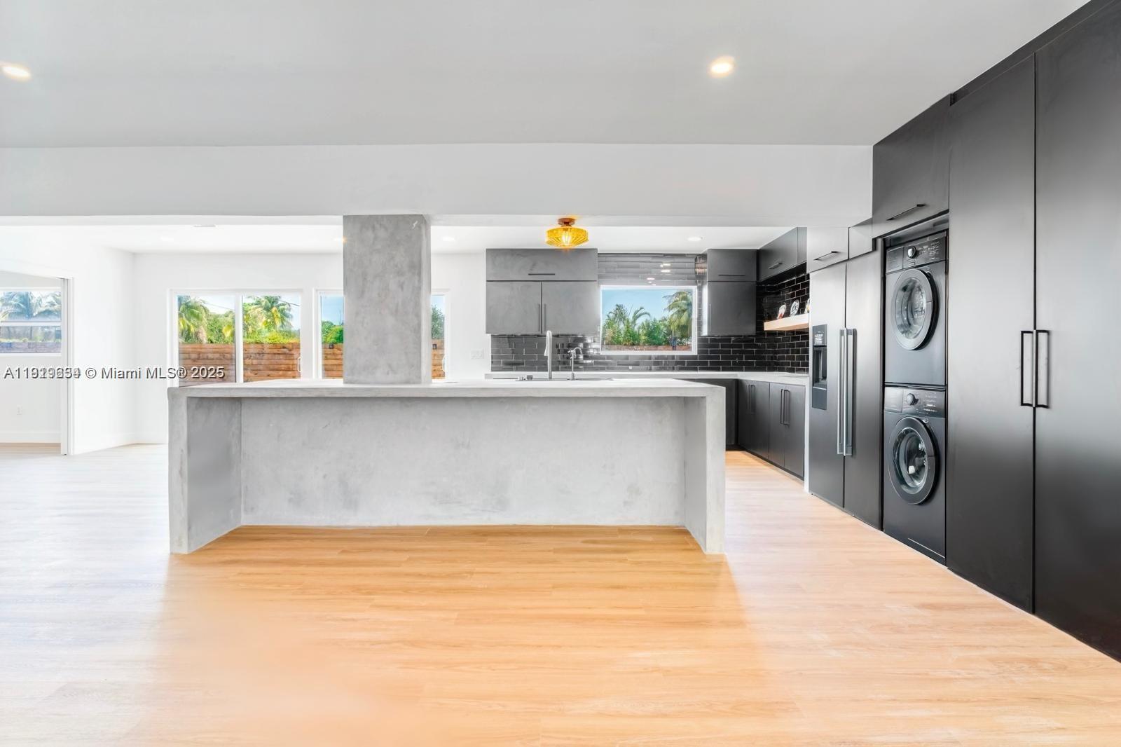 A modern kitchen features a concrete island, black cabinetry, and a stacked washer and dryer — 19661 NE 10th Pl, Miami FL 33179, MIAMI, FL