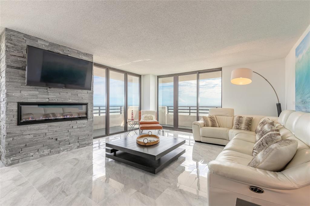 A modern living room features a stone fireplace, a white leather sofa, and floor-to-ceiling windows — 1420 N ATLANTIC AVENUE UNIT 601, DAYTONA BEACH, FL