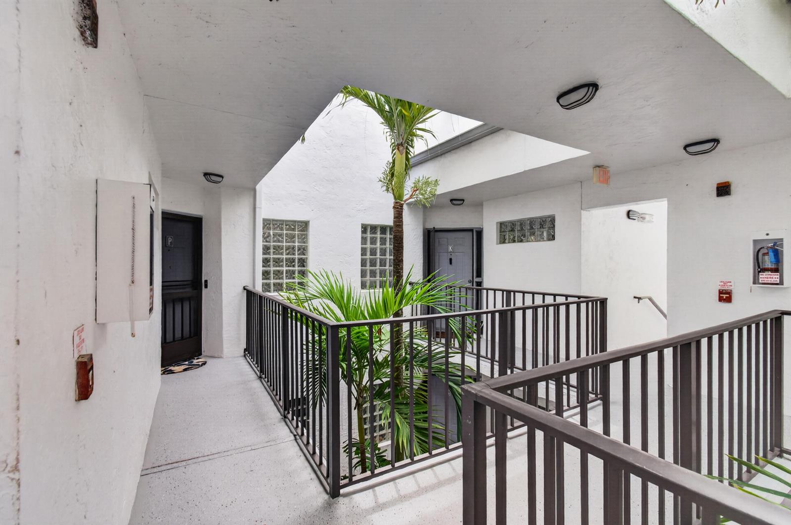 An open-air apartment building walkway features a central palm tree and dark metal railings — 5450 Verona Drive # P, Boynton Beach FL 33437, BOYNTON BEACH, FL