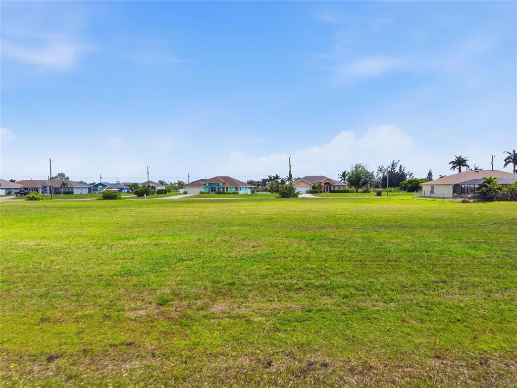 A wide, grassy vacant lot sits in front of several suburban houses under a blue sky — 3008 TROPICANA PARKWAY W, CAPE CORAL, FL