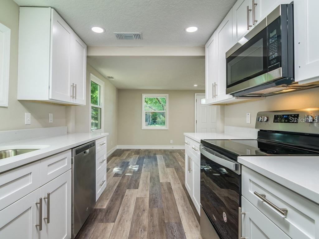 A galley kitchen with white cabinets, stainless steel appliances, and wood-look flooring leads to a room — 7225 33RD STREET N, ST PETERSBURG, FL