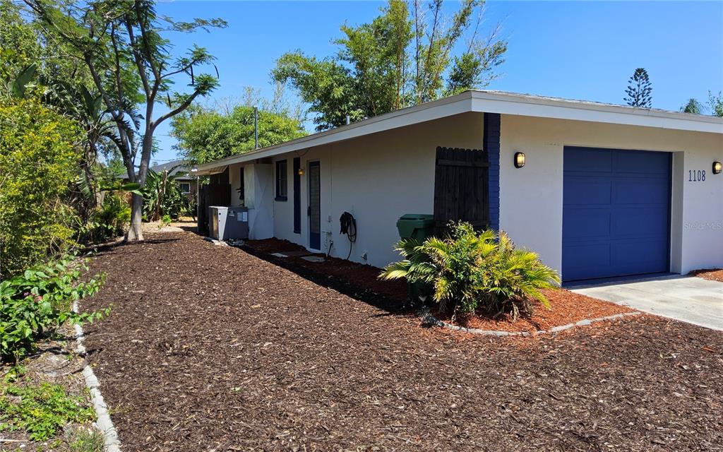 A side view of a house with a blue garage door and dark mulch landscaping — 1108 AMPLE AVENUE, PORT CHARLOTTE, FL
