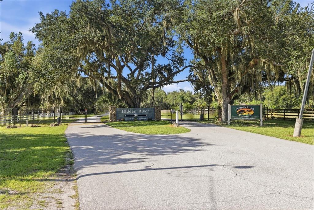 A gated entrance to a residential community features large oak trees and a stone sign — 3810 HIDDEN RIVER ROAD, SARASOTA, FL