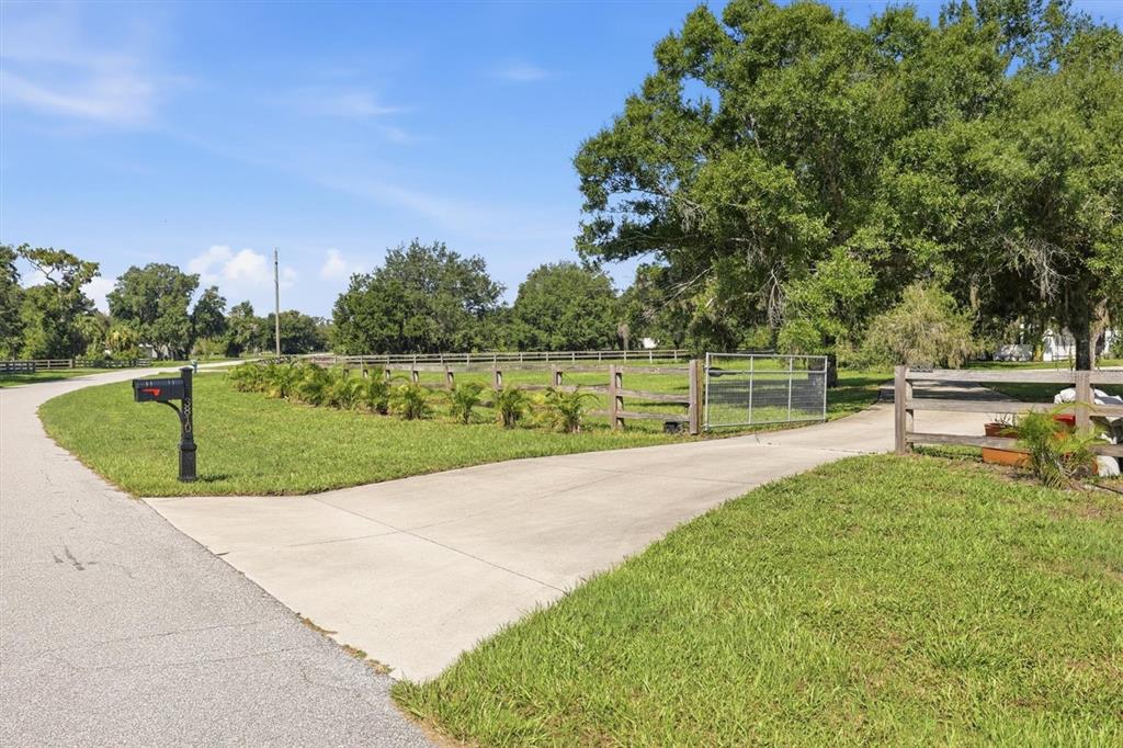 A concrete driveway leads through a wooden fence gate into a grassy, tree-lined property — 3810 HIDDEN RIVER ROAD, SARASOTA, FL