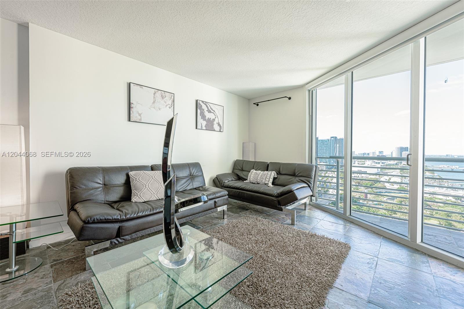 A living room features two dark leather sofas, a glass coffee table, and balcony views — 325 S Biscayne Blvd # 3624, Miami FL 33131, MIAMI, FL