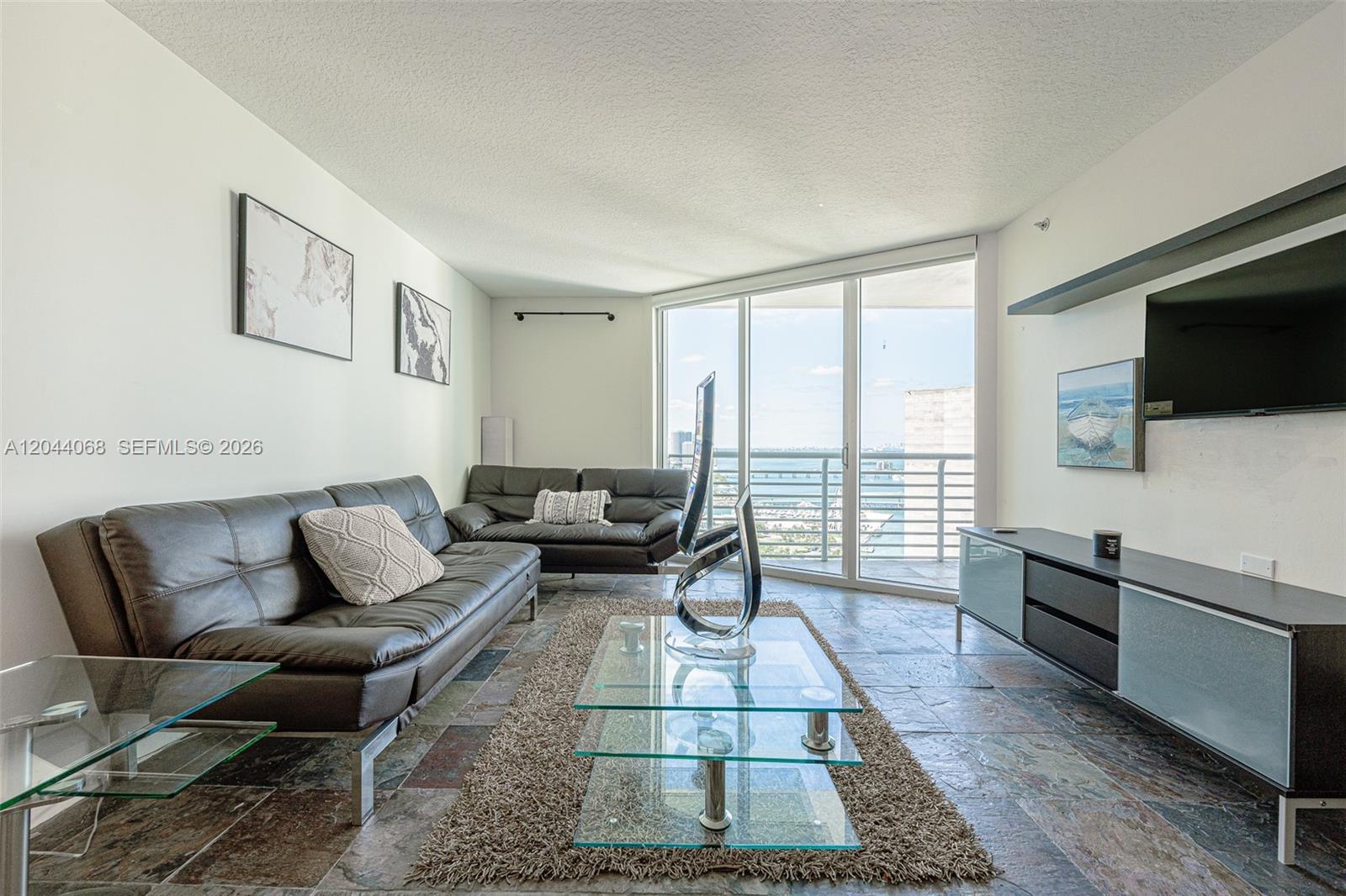 A living room features dark leather sofas, a glass coffee table, and a balcony view — 325 S Biscayne Blvd # 3624, Miami FL 33131, MIAMI, FL