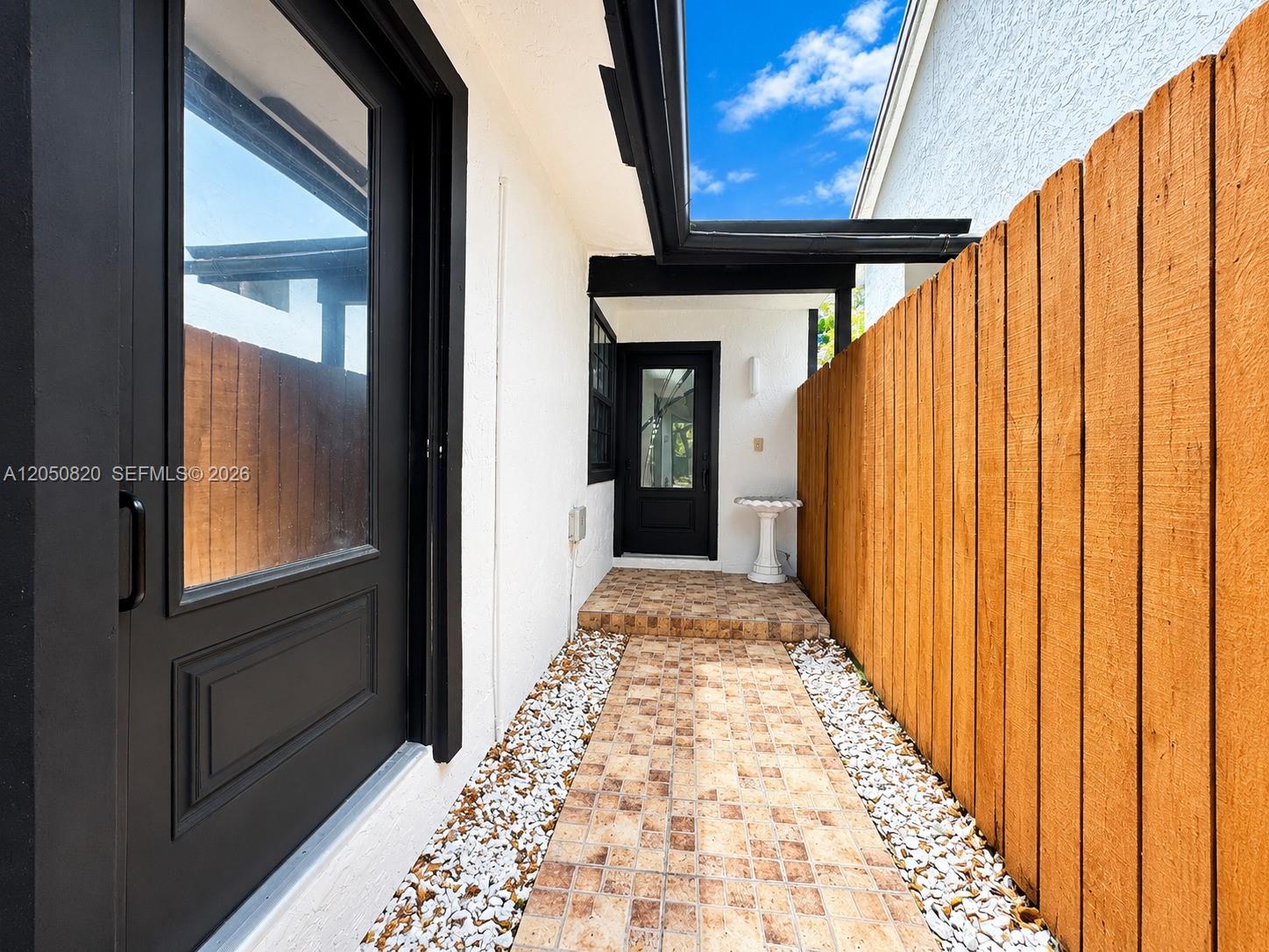 A tiled walkway leads to a black door between a white wall and a wooden fence — 10626 SW 148th Ave Dr, Miami FL 33196, MIAMI, FL