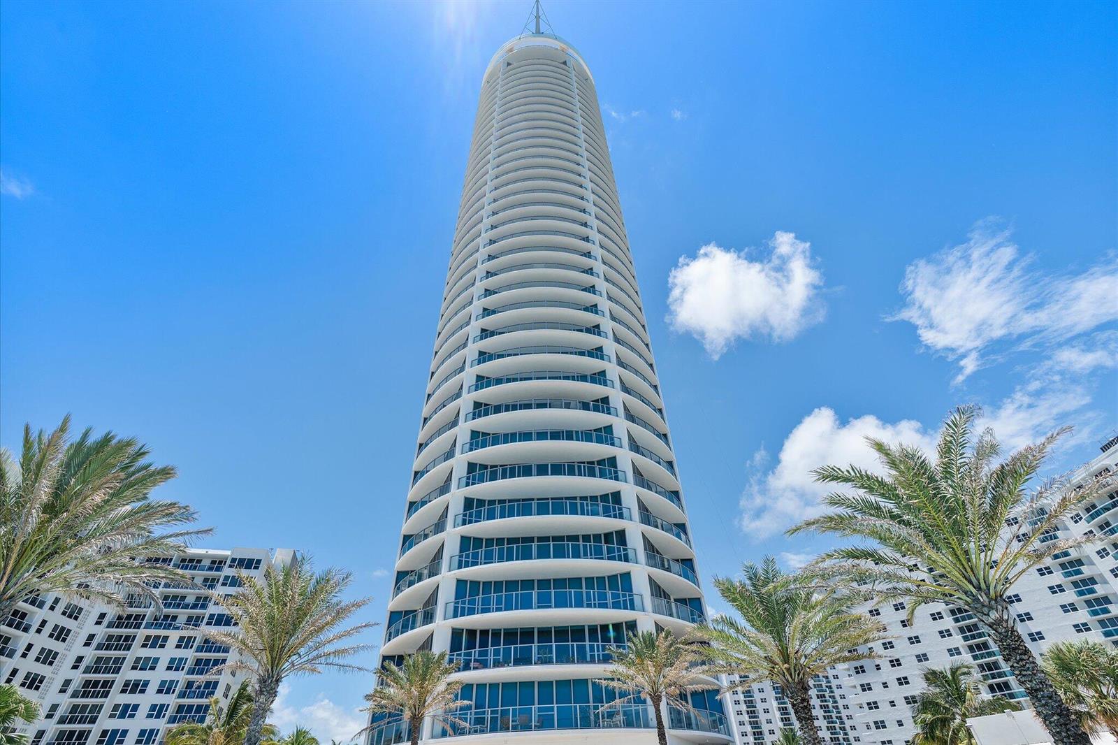 A low-angle view of a tall, cylindrical residential skyscraper surrounded by palm trees — 3101 S Ocean Drive # 2707, Hollywood FL 33019, HOLLYWOOD, FL