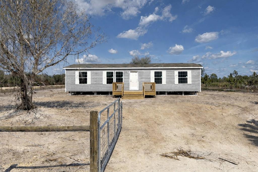 A single-story manufactured home with gray siding sits on a dirt lot behind a gate — 15983 NW 272ND STREET, OKEECHOBEE, FL