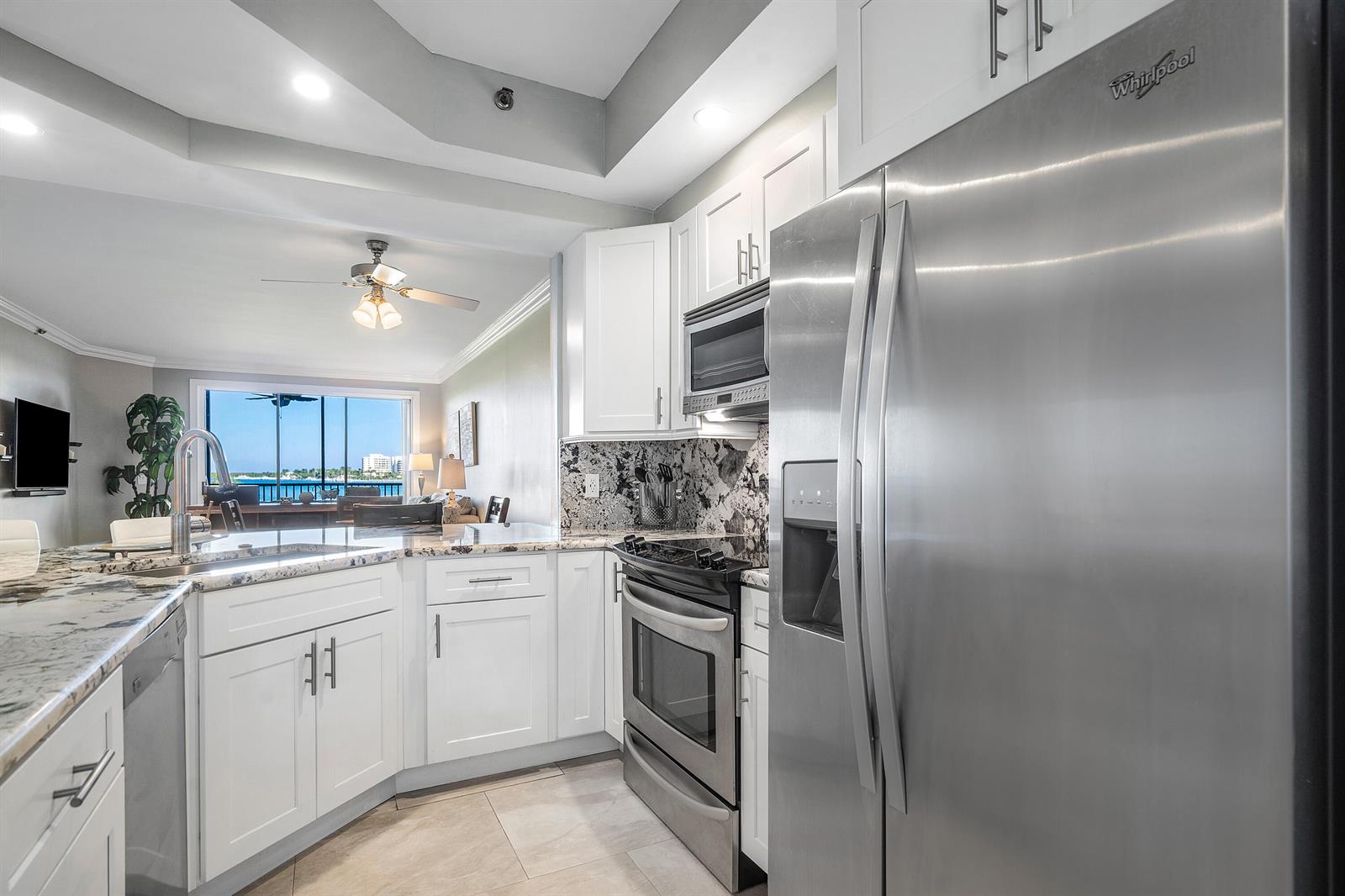 A modern kitchen features white cabinets, granite countertops, and stainless steel appliances overlooking a living area — 200 Waterway Road # 204, Tequesta FL 33469, TEQUESTA, FL
