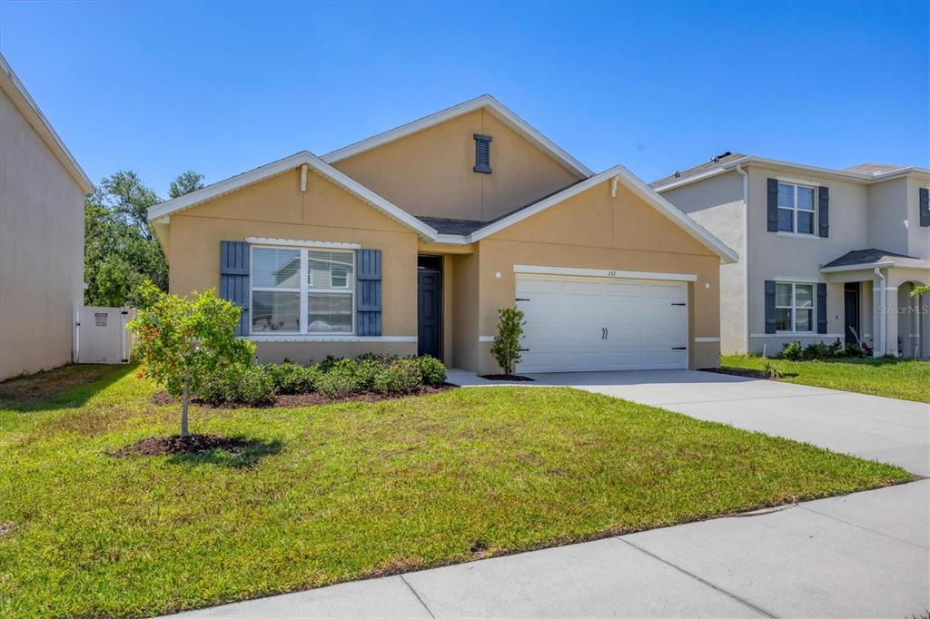 A tan single-story house with a two-car garage and blue shutters under a clear sky — 153 TIERRA VERDE WAY, BRADENTON, FL