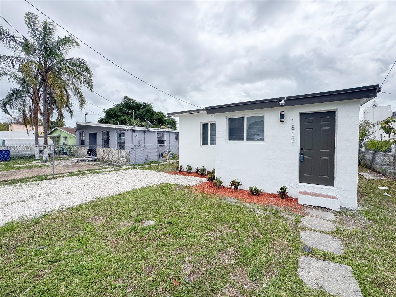 A white single-story house with a dark door and gravel driveway sits under a cloudy sky — 1822 NW 66th St, Miami FL 33147, MIAMI, FL