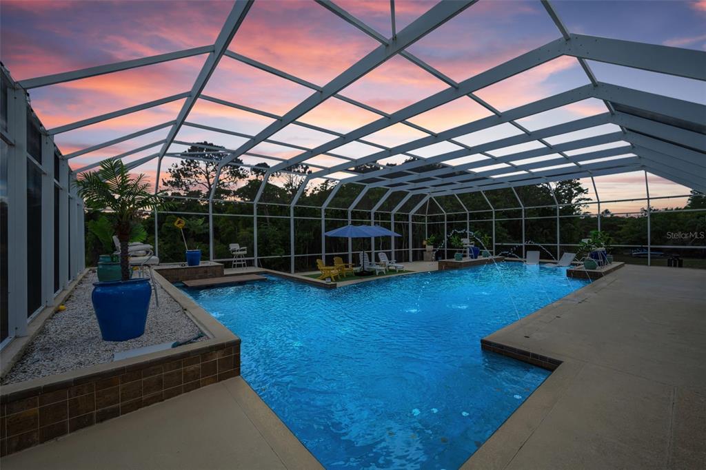 A screened-in swimming pool with fountains and patio furniture sits under a sunset sky — 11046 LAKEVIEW DRIVE, NEW PORT RICHEY, FL
