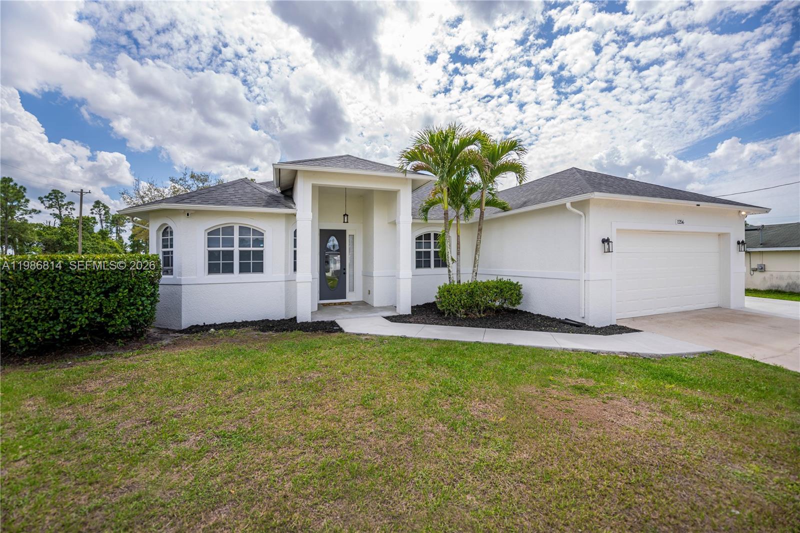 A white single-story house features a covered entryway, a two-car garage, and palm trees — 1256 E Edna St., Lehigh Acres FL 33974, LEHIGH ACRES, FL