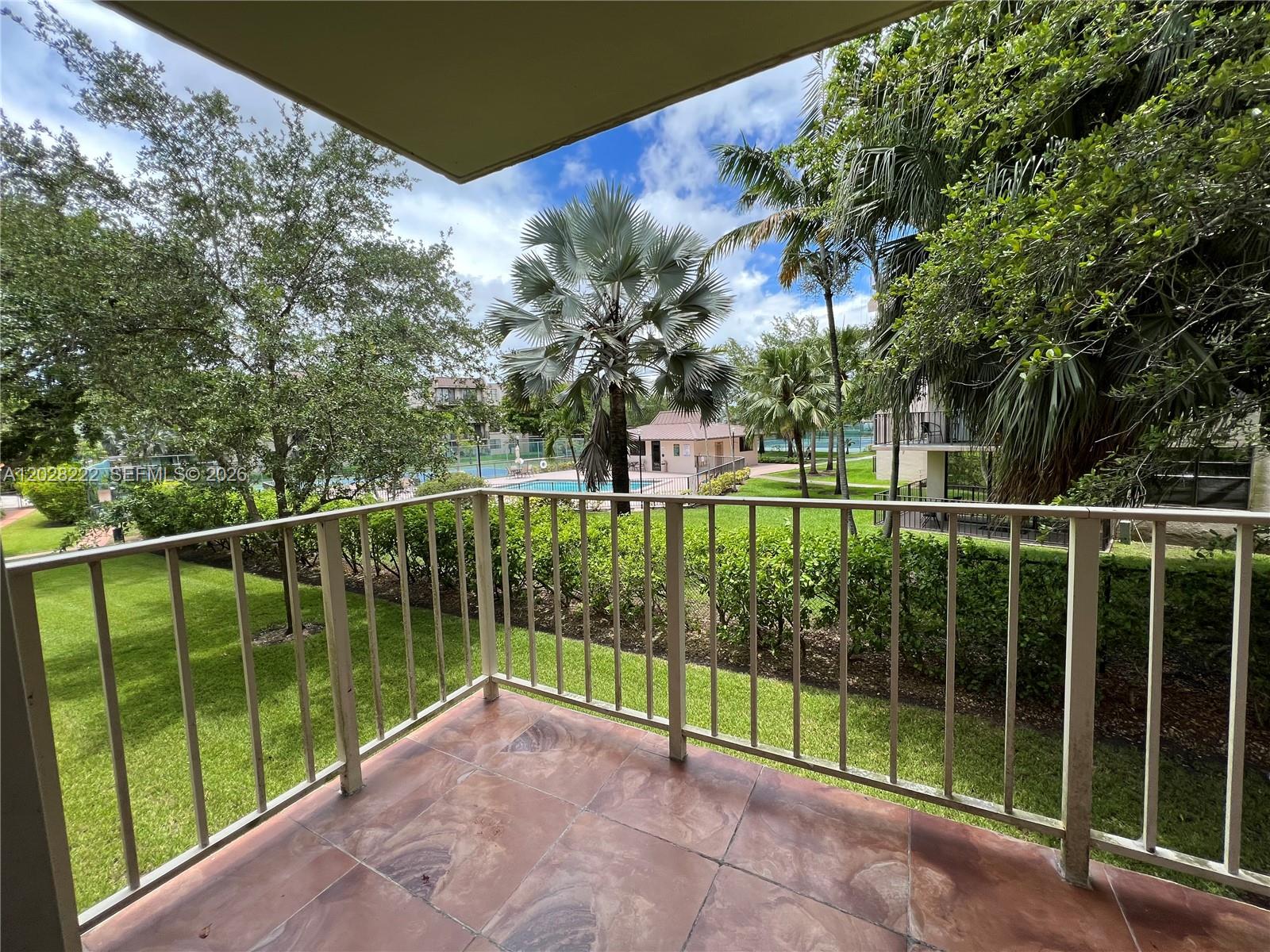A balcony with tiled flooring overlooks a grassy courtyard with a swimming pool and trees — 13100 SW 92nd Ave # 212, Miami FL 33176, MIAMI, FL