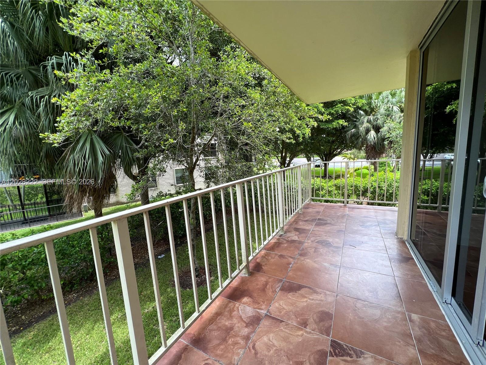 An outdoor balcony features brown tiled flooring, a white metal railing, and views of trees — 13100 SW 92nd Ave # 212, Miami FL 33176, MIAMI, FL