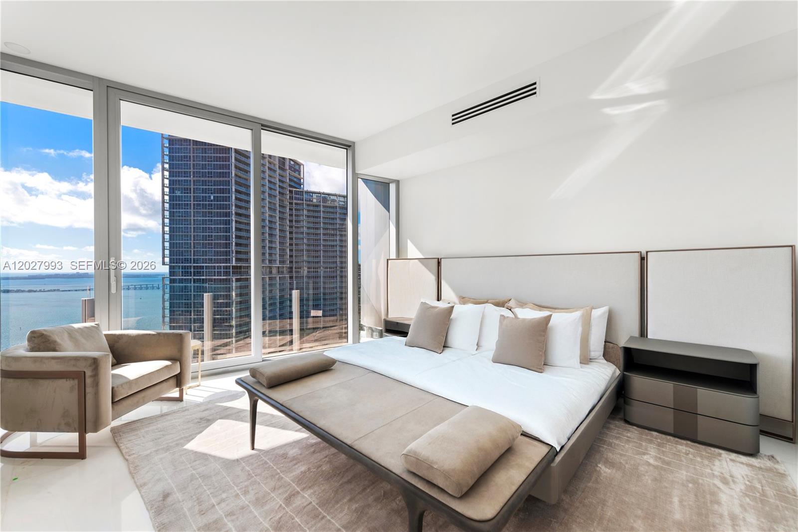 A modern bedroom features a large bed, a beige armchair, and floor-to-ceiling windows overlooking water — 300 Biscayne Boulevard Way # 3905W, Miami FL 33131, MIAMI, FL