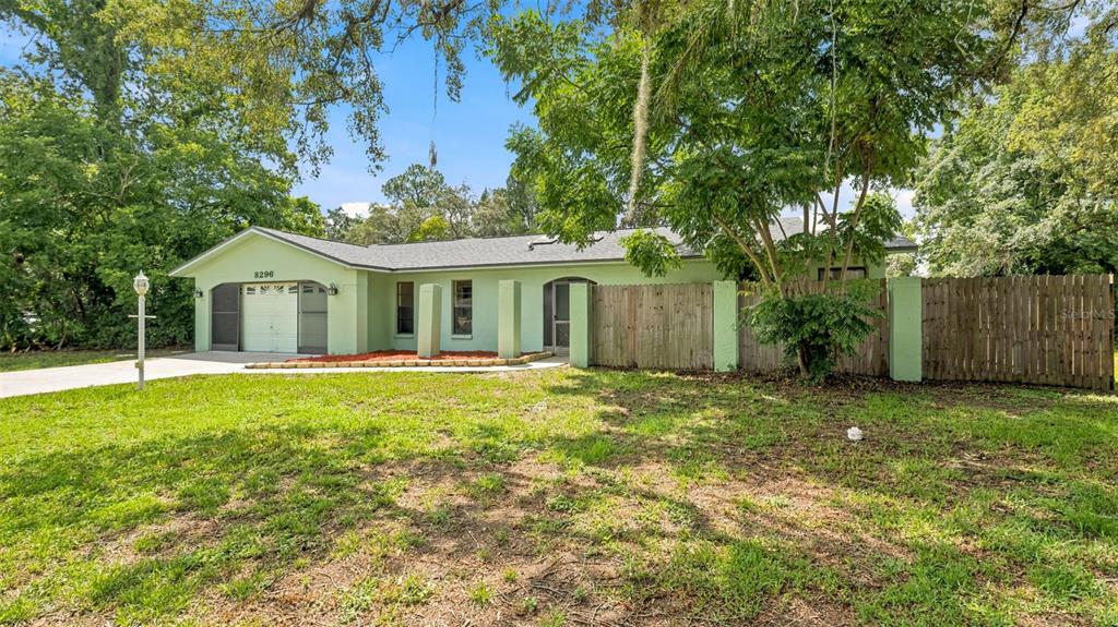 A light green single-story house features a garage, a wooden fence, and mature trees — 8296 GALLUP ROAD, SPRING HILL, FL