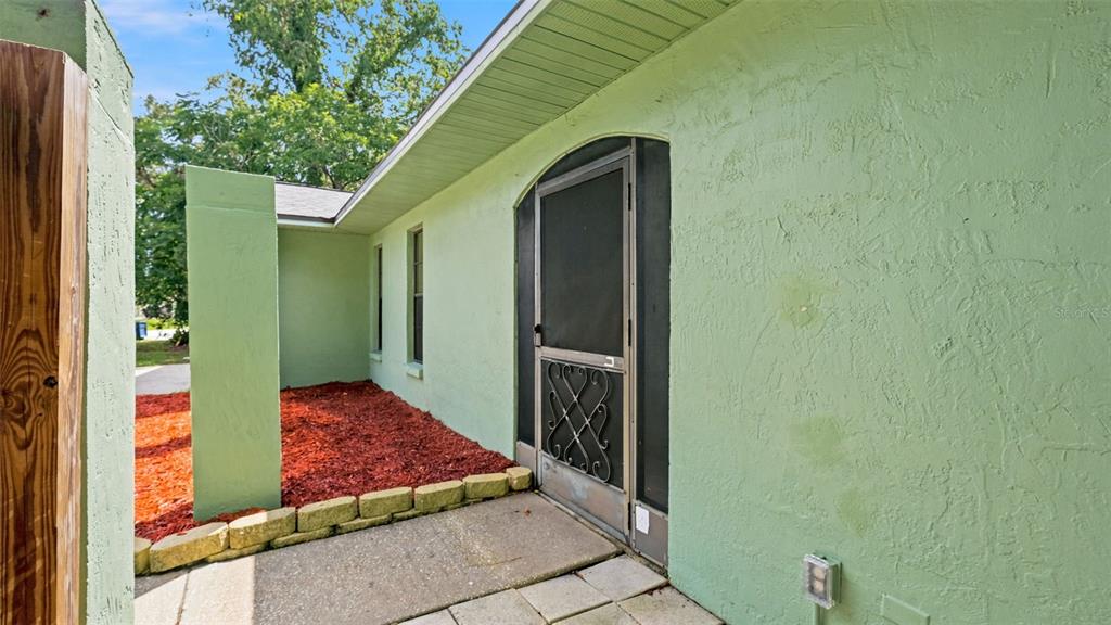 A light green stucco exterior wall features a screen door and a small garden bed — 8296 GALLUP ROAD, SPRING HILL, FL