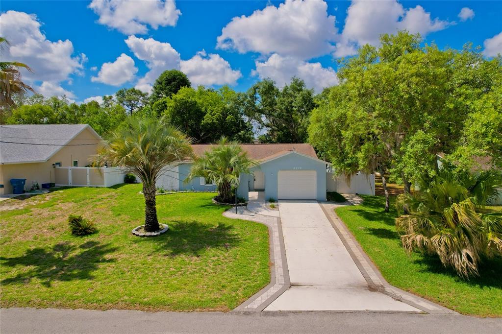 A light blue single-story house features a concrete driveway, palm trees, and a grassy lawn — 8218 PLACID STREET, SPRING HILL, FL