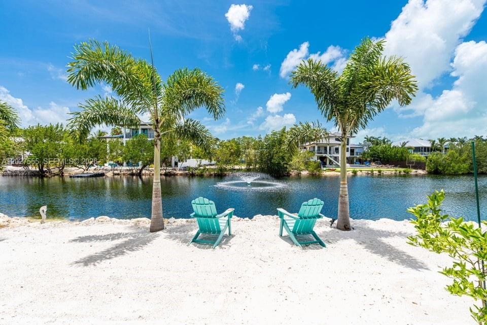 Two turquoise Adirondack chairs sit on a sandy beach overlooking a pond with a fountain — KEY LARGO, FL