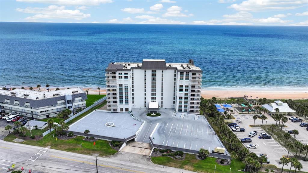 An aerial view shows a multi-story beachfront condominium building with a parking lot and ocean — 1575 OCEAN SHORE BOULEVARD UNIT 805, ORMOND BEACH, FL