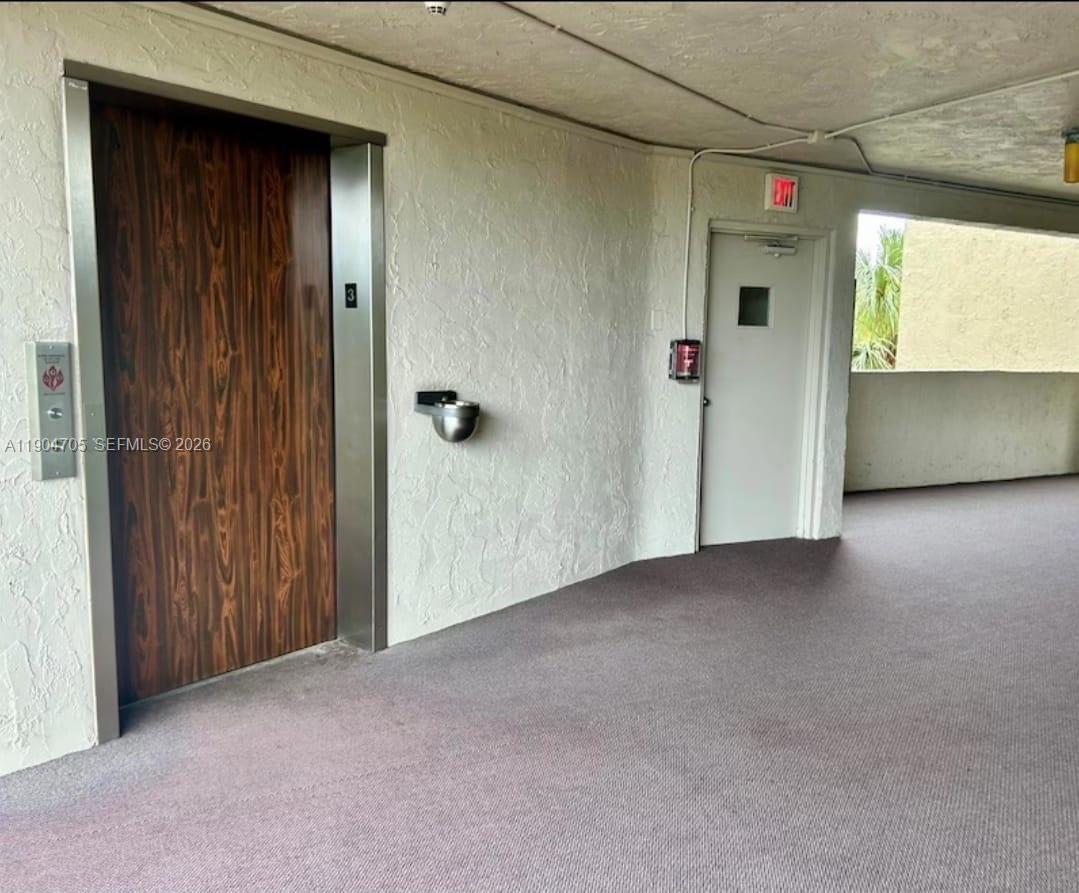 A hallway features an elevator with wood-grain doors, a white exit door, and carpeted flooring — 3910 Inverrary Blvd # B, Lauderhill FL 33319, LAUDERHILL, FL