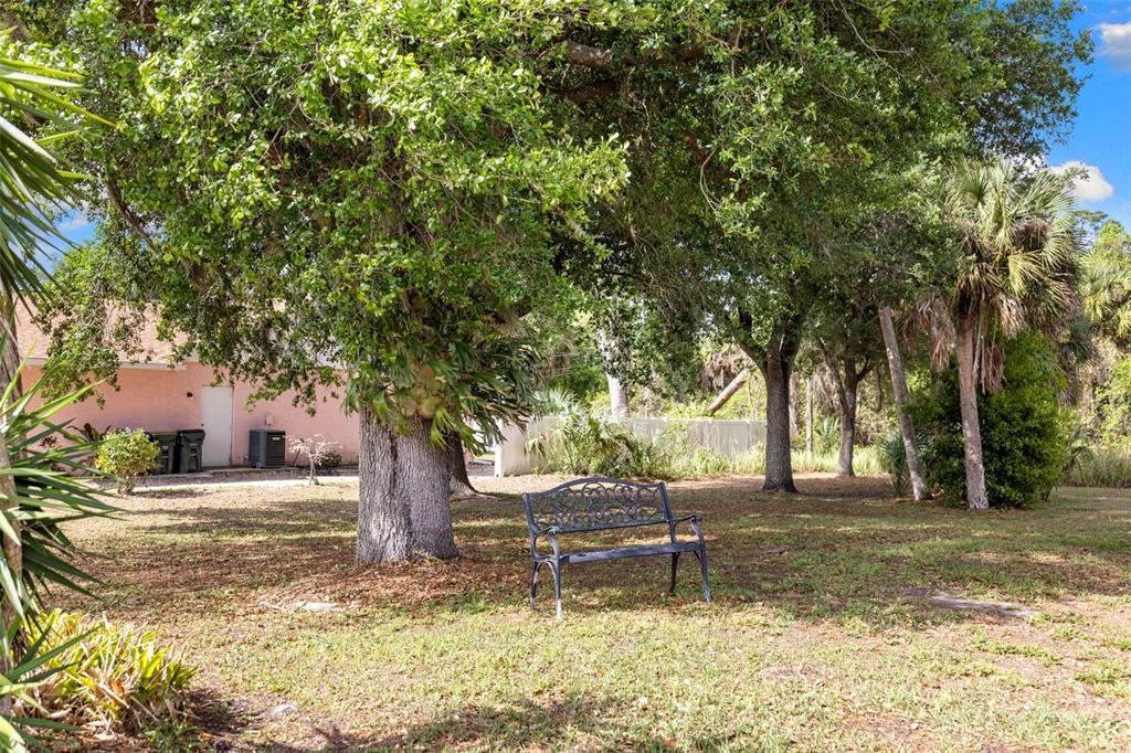 A black metal bench sits on a grassy lawn beneath large trees near a house — 4607 ATWATER DRIVE, NORTH PORT, FL