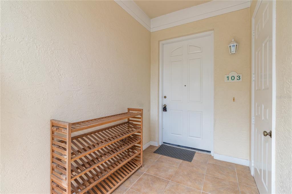 An entryway features a wooden shoe rack, tiled flooring, and a white front door labeled 103 — 3705 54TH DRIVE W UNIT 103, BRADENTON, FL